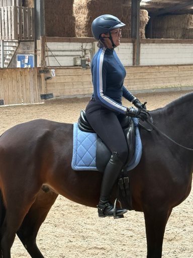 Reiterin auf einem dunklen Pferd in der Reithalle in der Seitenansicht. Die Reiterin zeigt das falsche Sitzmuster "Einknicken in der Hüfte"